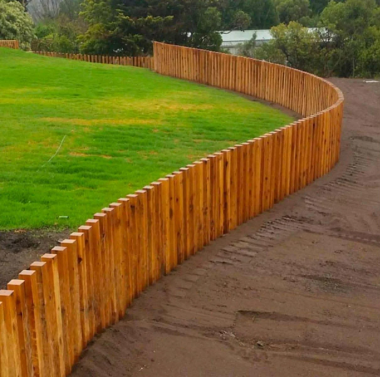 A newly built wooden fence curves along the edge of a grassy lawn, dividing it from a dirt path. Trees and bushes are visible in the background.