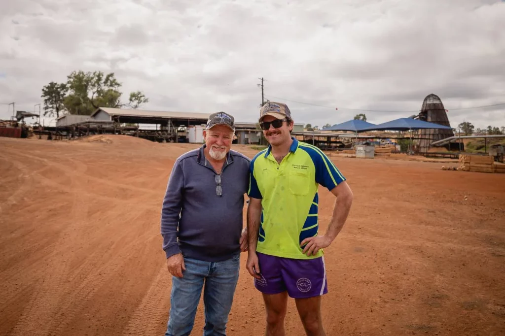 Two men stand together on a dirt lot, smiling at the camera. One wears a blue sweater and jeans, the other a bright green shirt and purple shorts. Sheds and farm buildings are visible in the background under a cloudy sky.