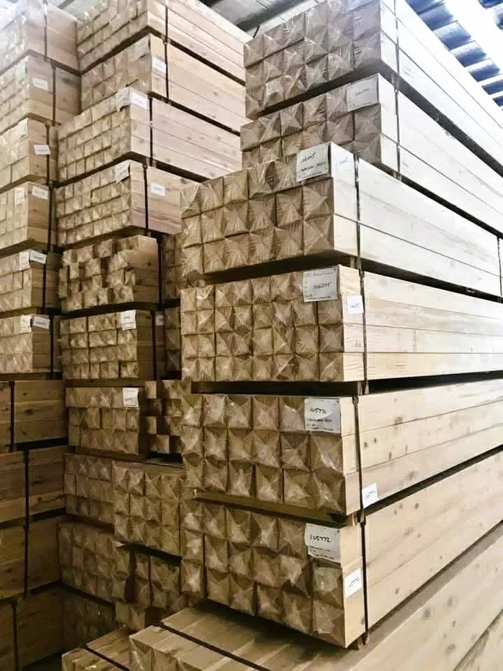 Stacks of neatly arranged wooden beams with decorative carved ends, stored indoors in a warehouse or lumberyard. Paper labels are attached to the ends of some beams for identification.