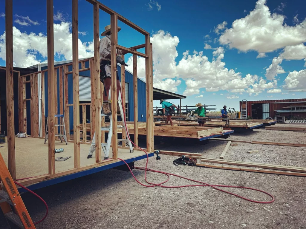 Workers build wooden frames for a structure under a bright, cloudy sky. One person stands on a ladder, while others work in the background. Construction materials and tools are scattered on the ground.