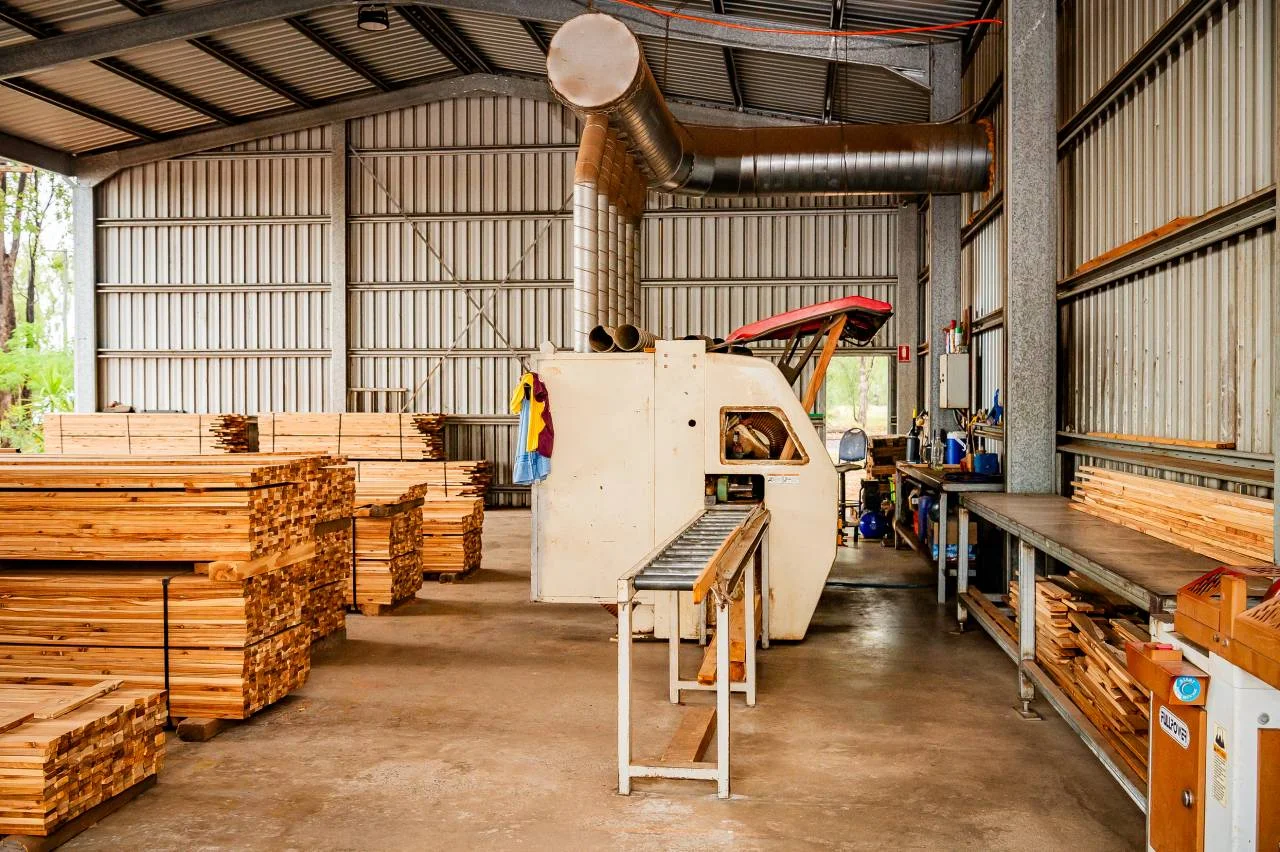 Stacks of cut timber are piled inside a metal shed. A woodworking machine with a conveyor belt sits in the center, surrounded by workbenches and tools, in a well-lit, organized workshop.